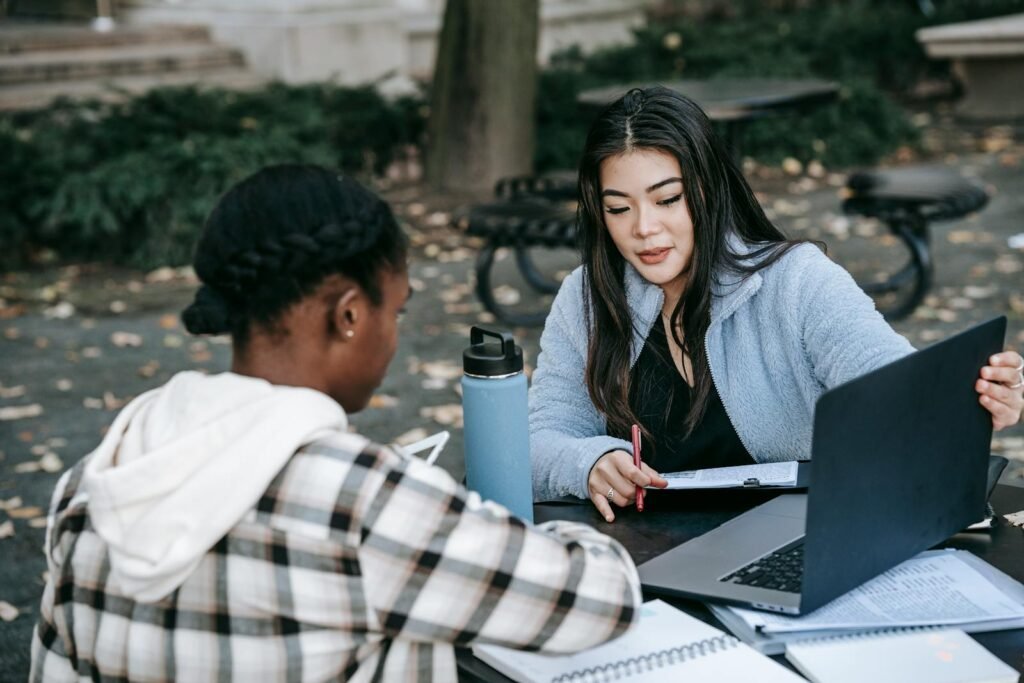 Two young women studying together outdoors on campus with laptops and notes.